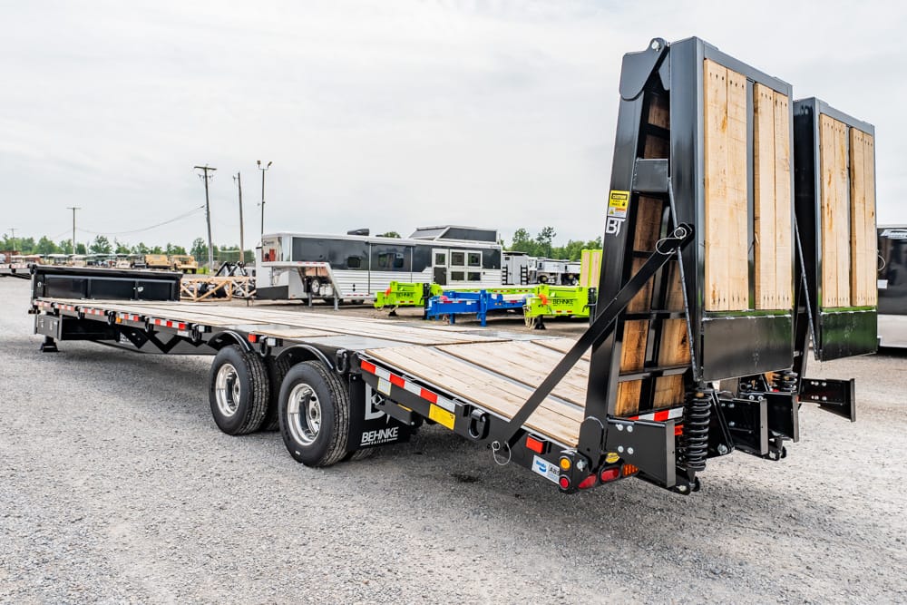 Flatbed trailer with loading ramp in a gravel lot, escorted by spare equipment and trailers in the background.