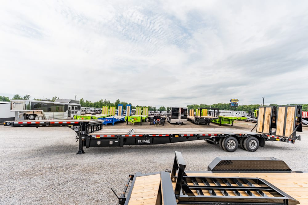 Long flatbed trailer with wooden ramps in a gravel lot, surrounded by colorful trailers in the background.