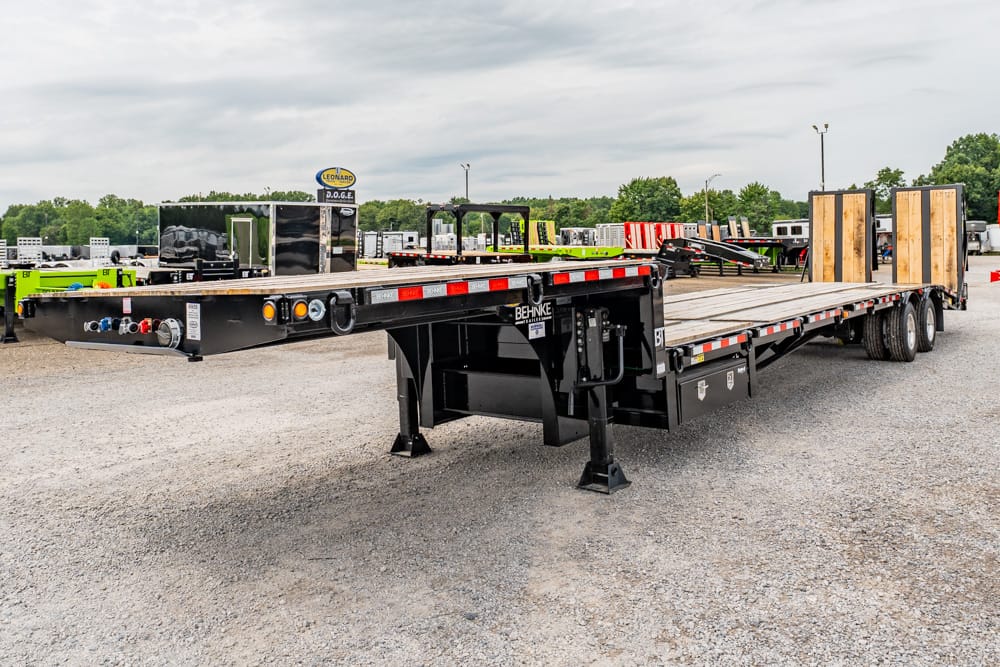 Long black flatbed trailer with wooden loading ramps in a gravel trailer yard, ready to haul heavy equipment.