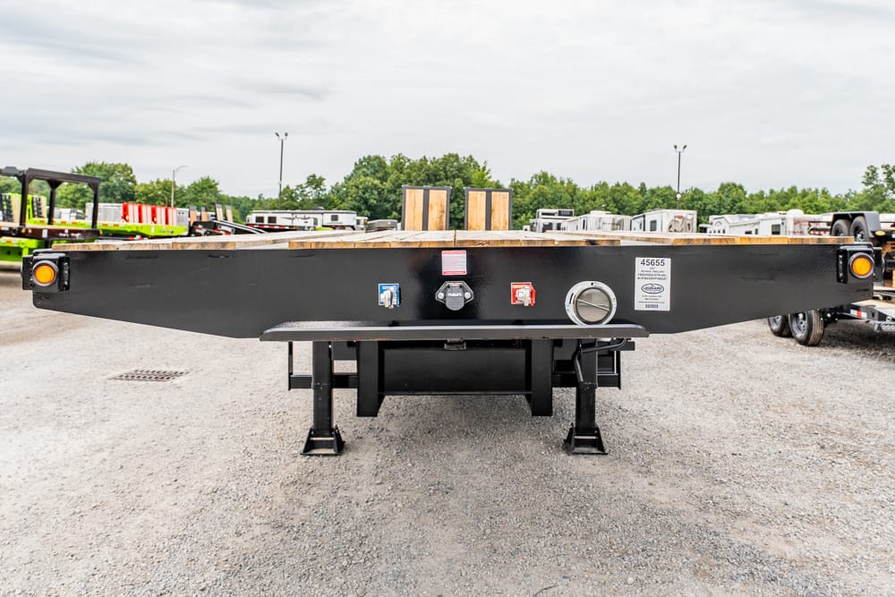 Rear view of a black flatbed trailer with a wooden deck, parked on gravel in a equipment yard, showing lights and control valves.