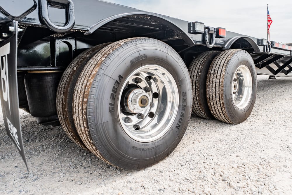Close-up of dual trailer wheels with chrome rims on a gravel lot; part of a flatbed trailer is visible with an American flag in the distance.