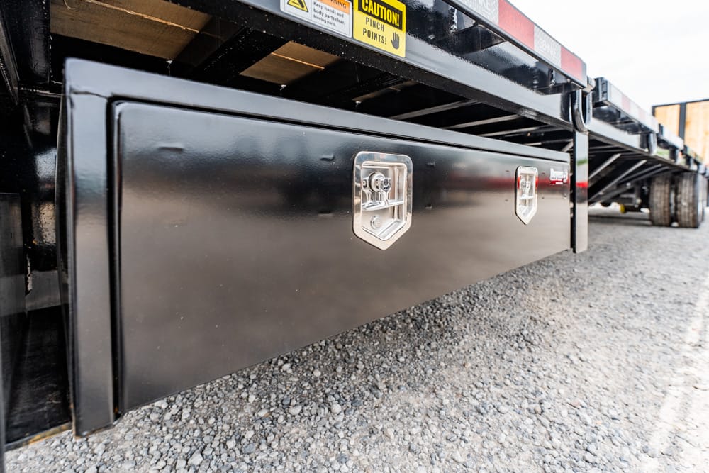 Low-angle shot of a black metal undercarriage toolbox mounted to a flatbed trailer, security latches visible, on gravel.