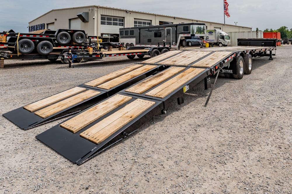 Long flatbed trailer with four wooden ramps extended on a gravel lot at a trucking yard, ready to load vehicles.