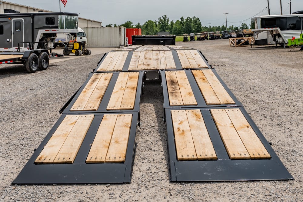 Long flatbed trailer ramp with wooden planks in a gravel lot, industrial buildings and parked trucks in the background