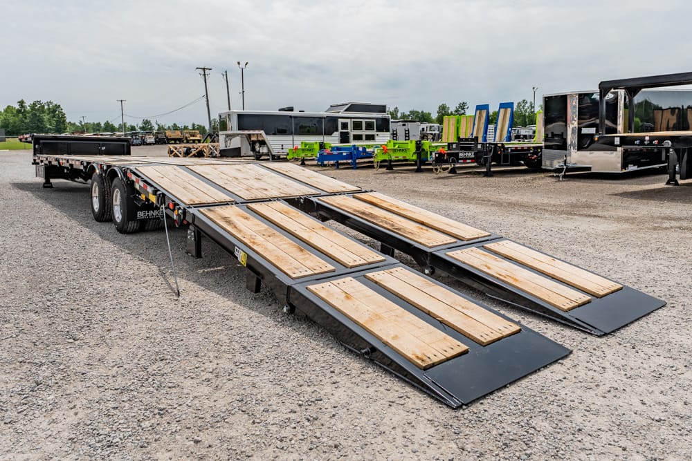 Flatbed trailer with four wooden loading ramps extended on a gravel lot, ready for vehicle loading, with equipment in the background.