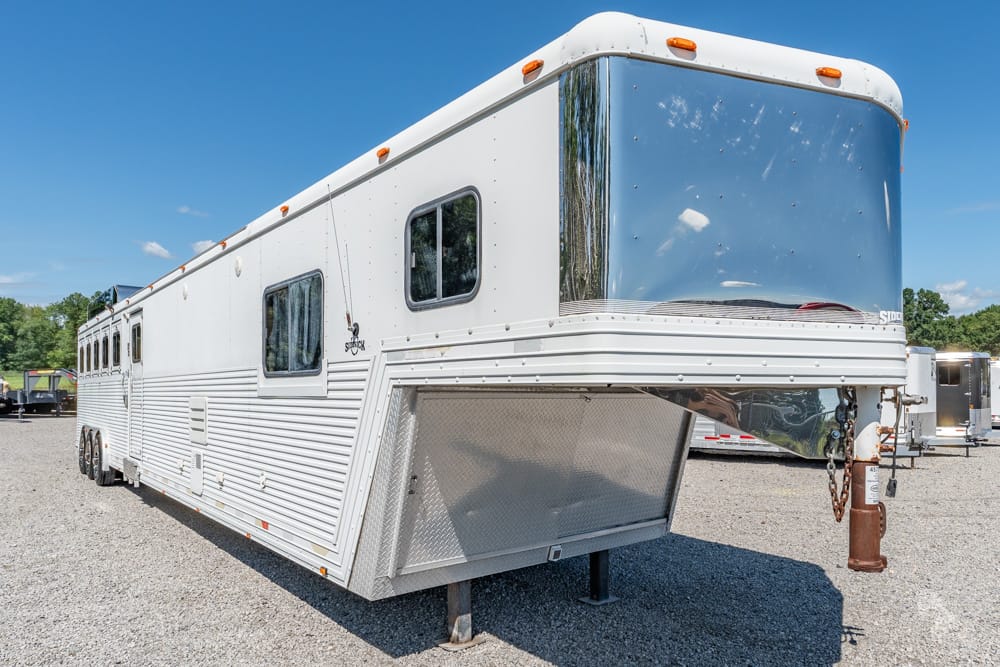 White gooseneck fifth-wheel horse trailer parked on a gravel lot under a clear blue sky, with chrome front and windows along the side