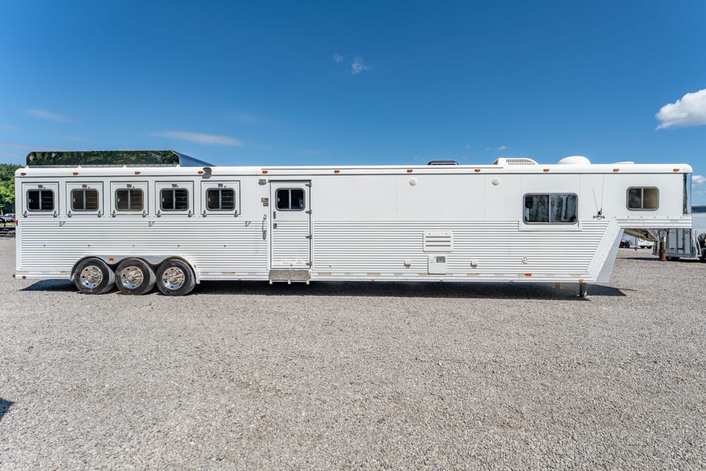 White horse trailer with three axles parked on a gravel lot under a clear blue sky, side view showing windows and doors.