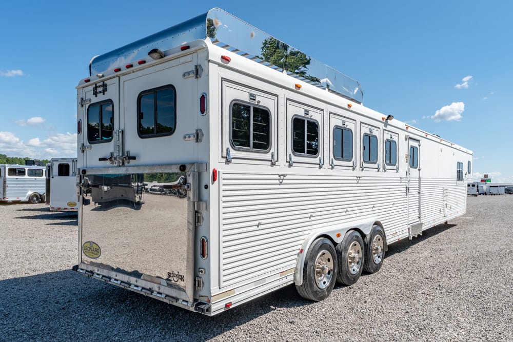 Large aluminum horse trailer with three axles and multiple windows parked on a gravel lot under a clear blue sky