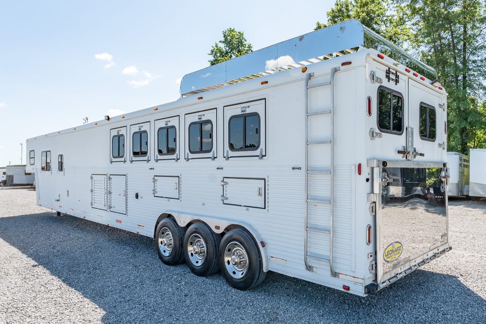 White enclosed horse trailer with multiple side windows and a rear ramp, parked on a gravel lot under a blue sky, trees in the background.