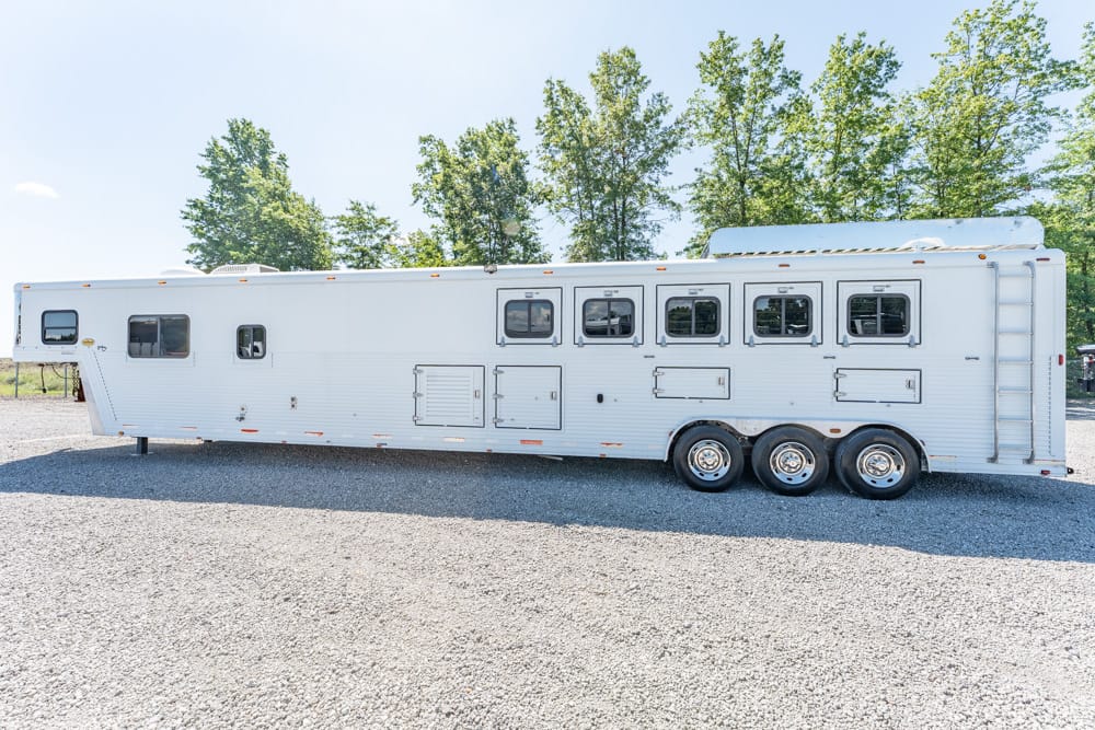 White long horse trailer with three axles and multiple windows parked on a gravel lot, with trees in the background.
