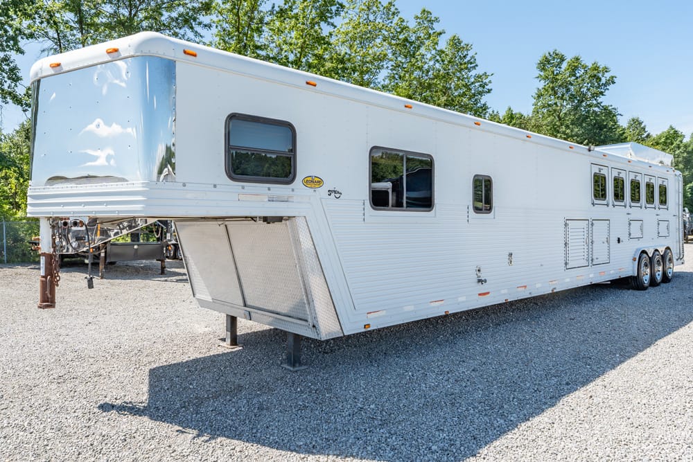 Long white gooseneck horse trailer with multiple windows parked on a gravel lot, raised front hitch and stabilizing leg visible under a clear blue sky.