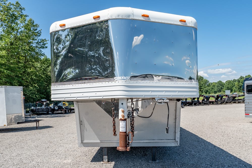 Front view of a large silver gooseneck trailer with a curved windshield, parked on a gravel lot with trees in the background.