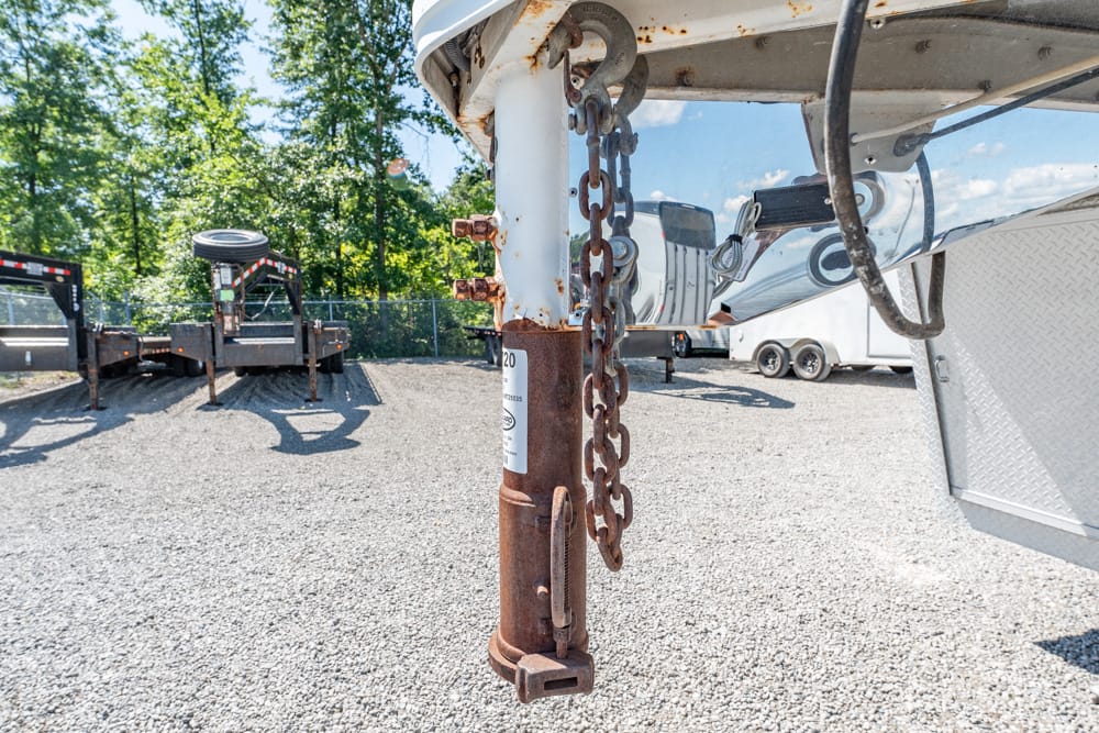 Close-up of a rusted trailer jack and chain on the hitch of a parked trailer in a gravel lot with other trailers in the background