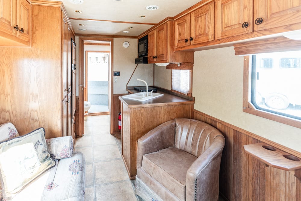 Wood-paneled RV kitchen with a sink island, overhead cabinets, and a leather armchair beside a window.