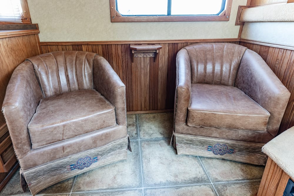 Two brown leather armchairs in a vintage wood-paneled RV interior with a tiled floor and windows above them, facing each other.