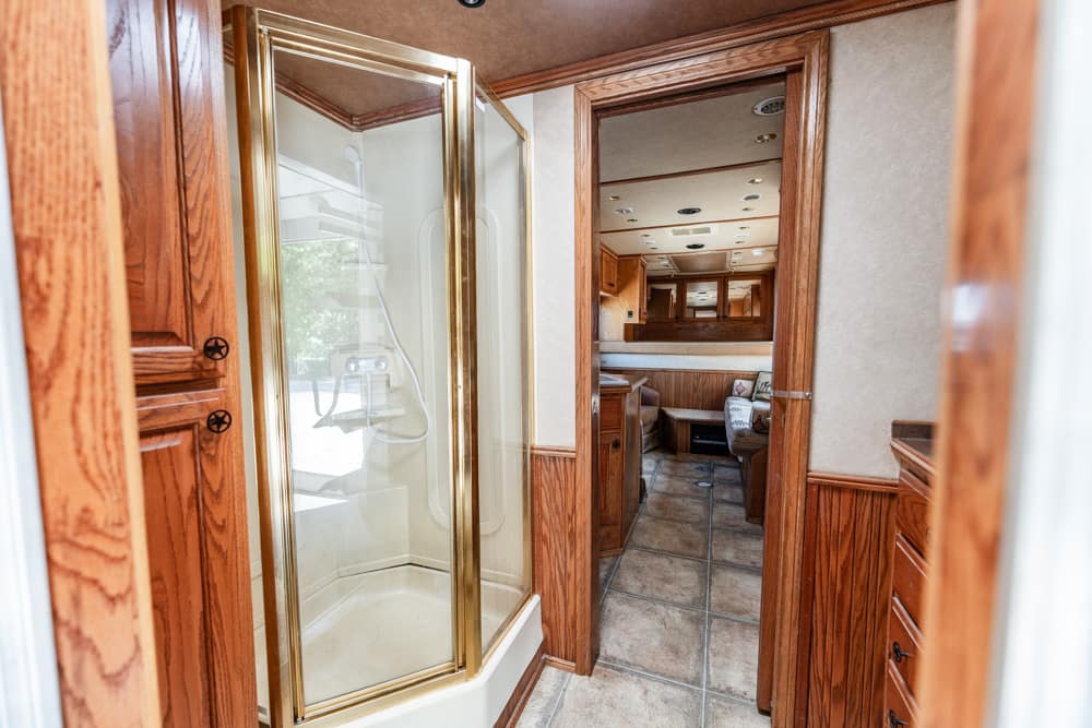 Glass-walled shower with gold trim in a wood-paneled RV bathroom, open door revealing a tile floor and cabinetry nearby, leading to a living area.