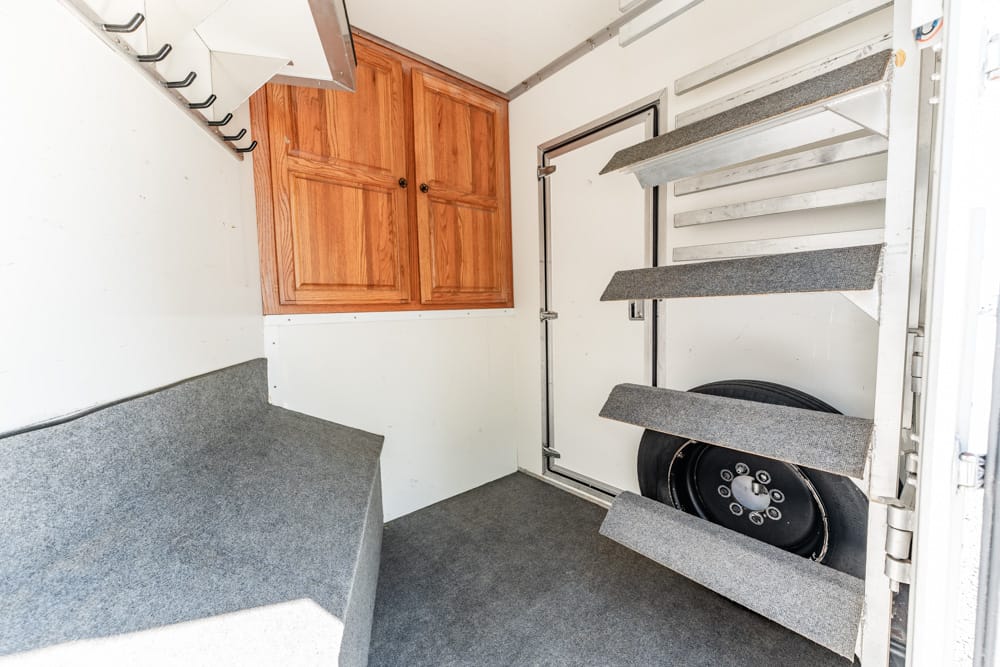 Interior of a utility trailer workshop with wooden cabinets, a gray padded bench, and carpeted stair steps near the door.