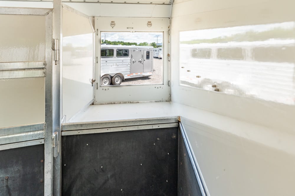 Interior of a light trailer looking out through a small window at a parked silver horse trailer in a gravel lot.