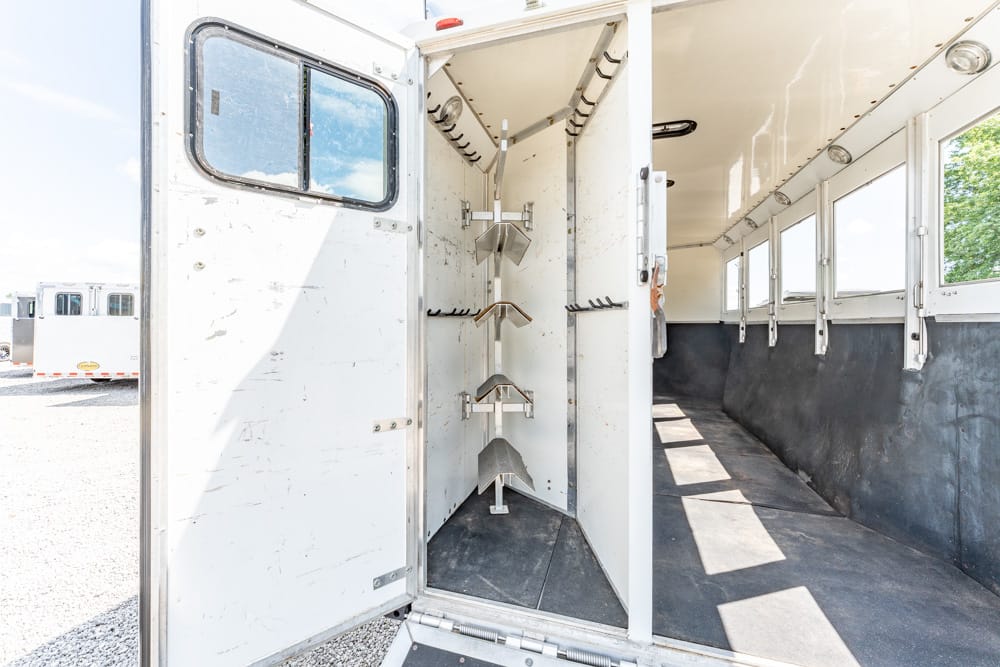 Inside of a white horse trailer showing a corner-mounted saddle rack system, an aisle with windows along the right wall, and a rubberized floor.