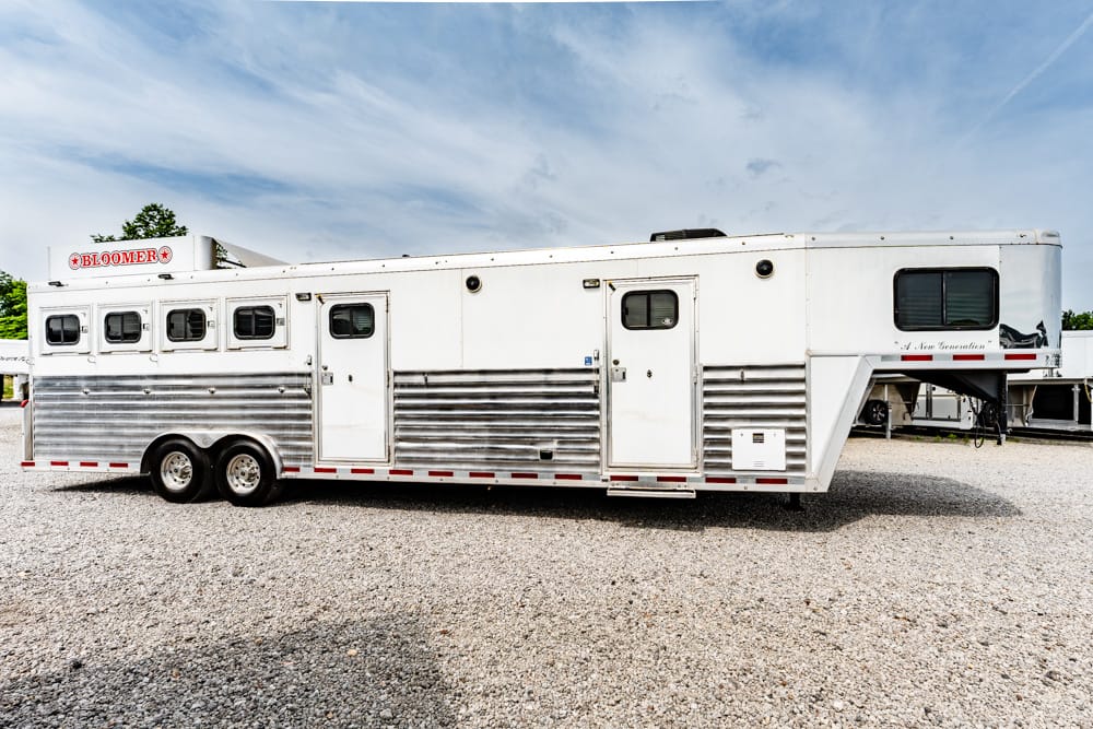 White Bloomer horse trailer with windows and dual rear wheels parked on a gravel lot.