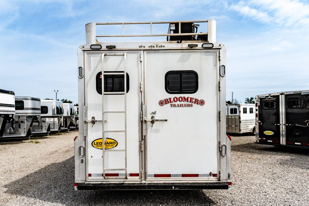 Rear view of a white Bloomer trailer with a built-in ladder on the left, a small rear window, and other trailers in a gravel lot.