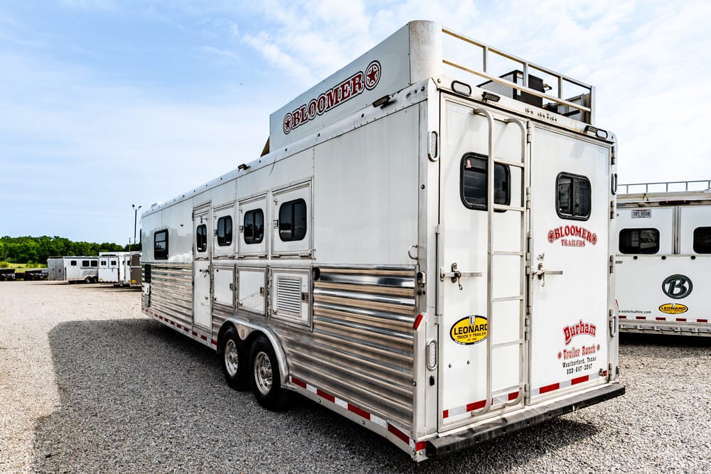 White Bloomer horse trailer with polished aluminum lower siding, parked on a gravel lot with other trailers in the background.