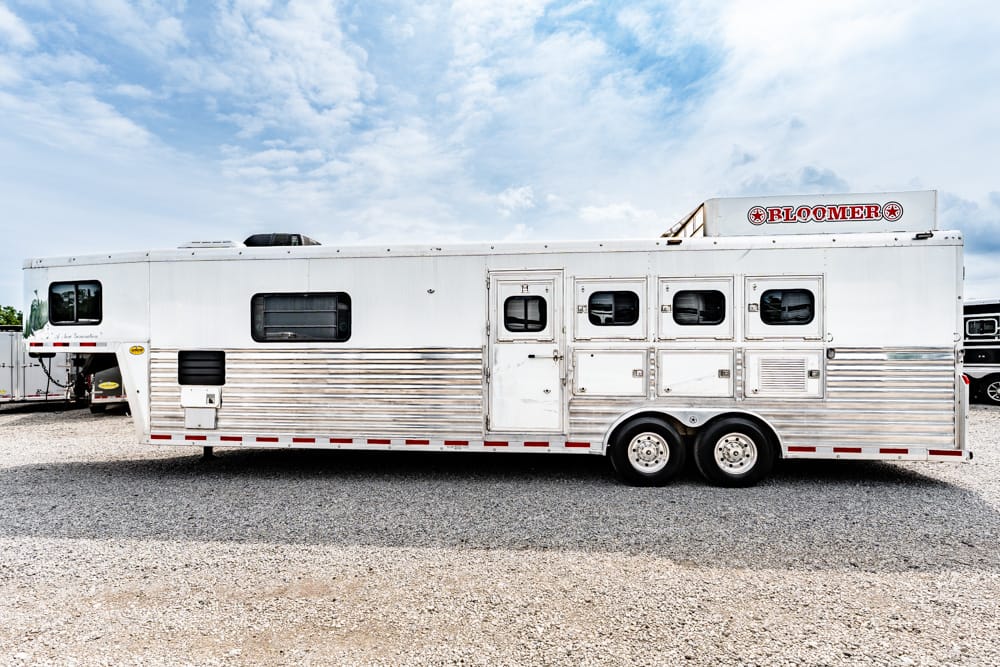 Long white-and-silver gooseneck horse trailer with multiple stalls and windows, parked on a gravel lot under a blue sky, Bloomers branding on top edge.