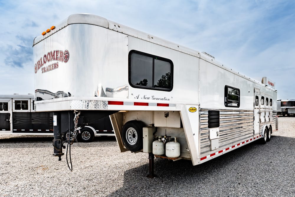 Silver Bloomer gooseneck horse trailer with propane tanks and a spare tire under the front overhang, parked on gravel.