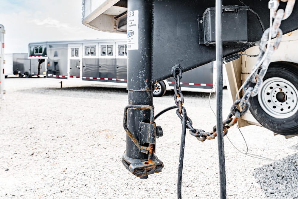 Close-up of a rusted trailer hitch and safety chains on a black hitch, with gravel ground and horse trailers in the background.