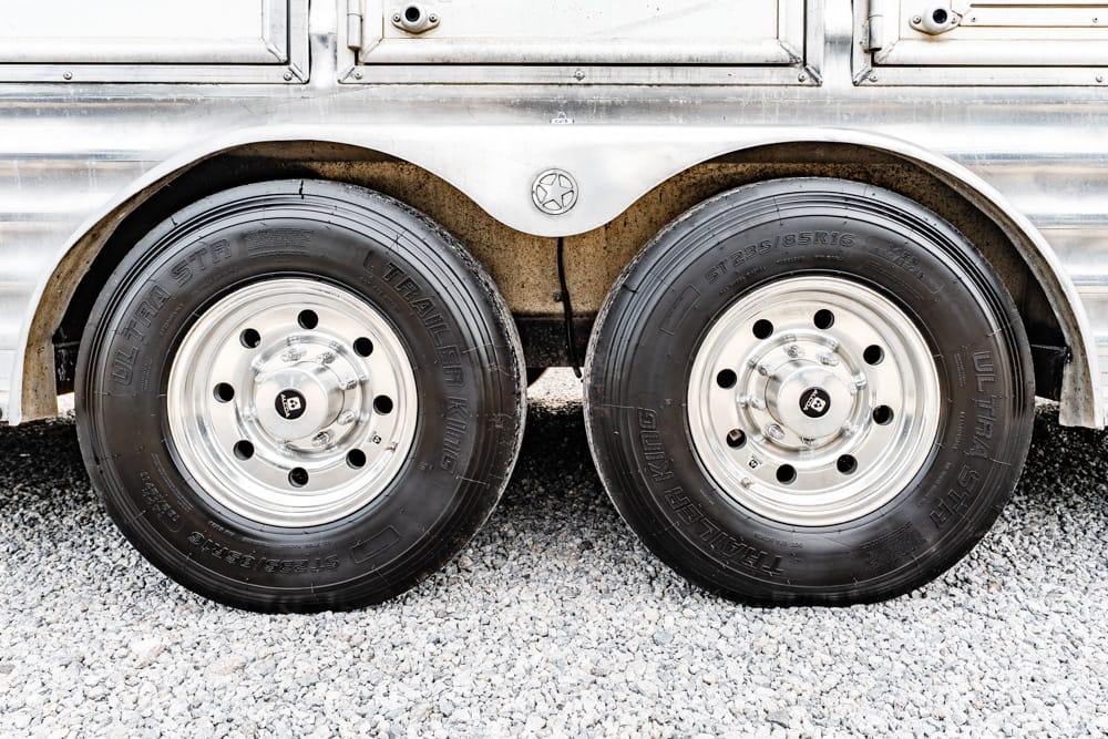 Dual trailer wheels with chrome rims on a silver trailer, resting on a gravel surface.