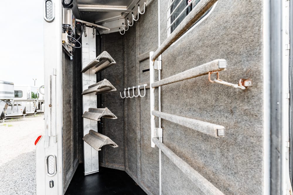 Interior of a horse trailer with wall-mounted saddle racks and hooks, gray carpeted panels, and narrow aisle inside the trailer