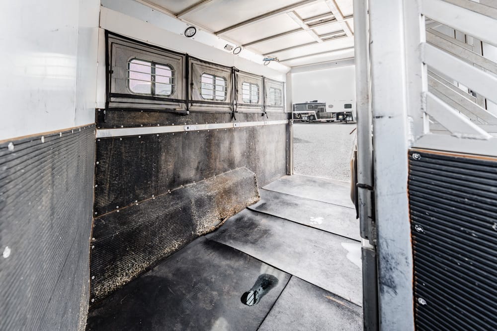 Interior of a metal livestock/trailer with rubber mats, side walls, and small windows along the top, leading outside to parked trucks.