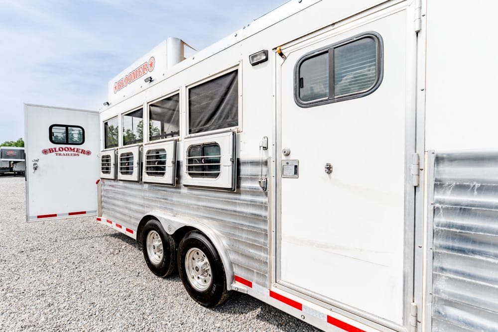 White Bloomer horse trailer with side windows and rear door open on a gravel lot.