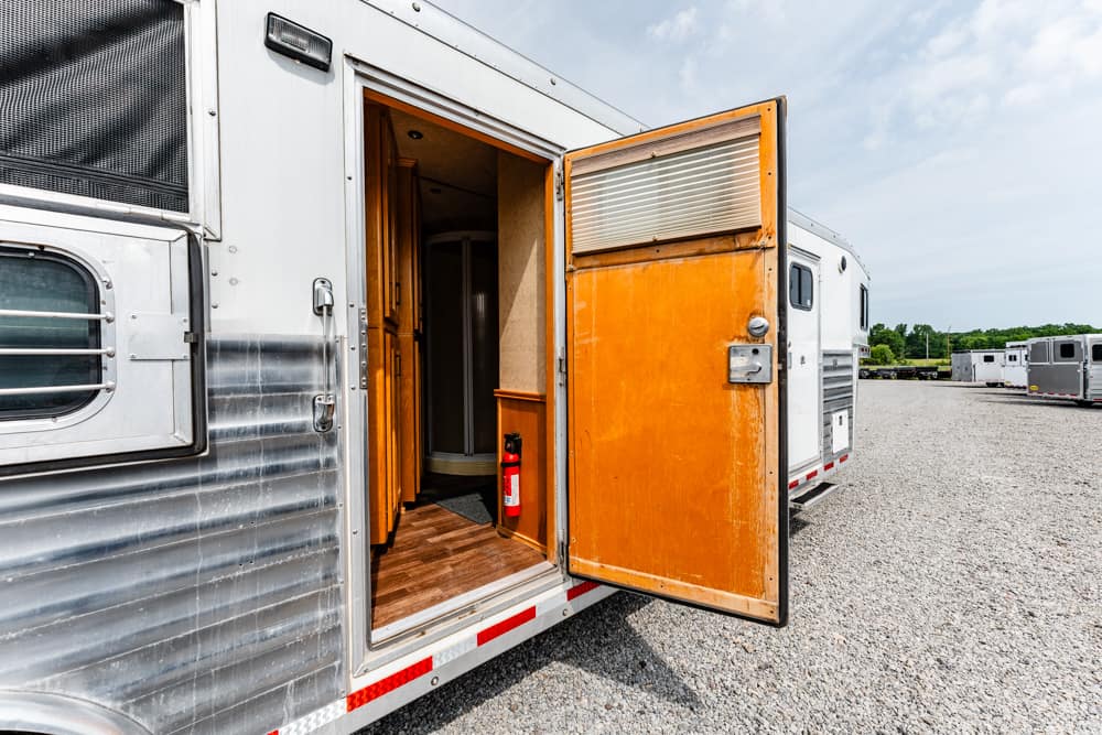 Open orange side door of a silver horse trailer; visible inside are wood panels and a red fire extinguisher, with a gravel lot and other trailers in the distance.