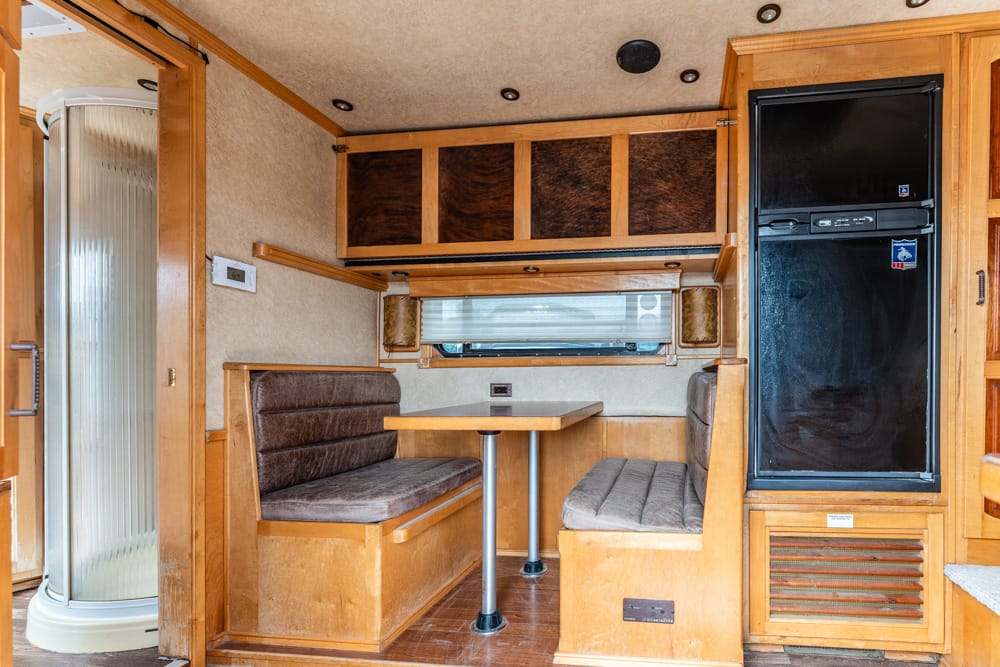 RV dining area with booth seating, a wooden table, window blinds, and overhead cabinets beside a tall black refrigerator league area open to left shower stall in the corner