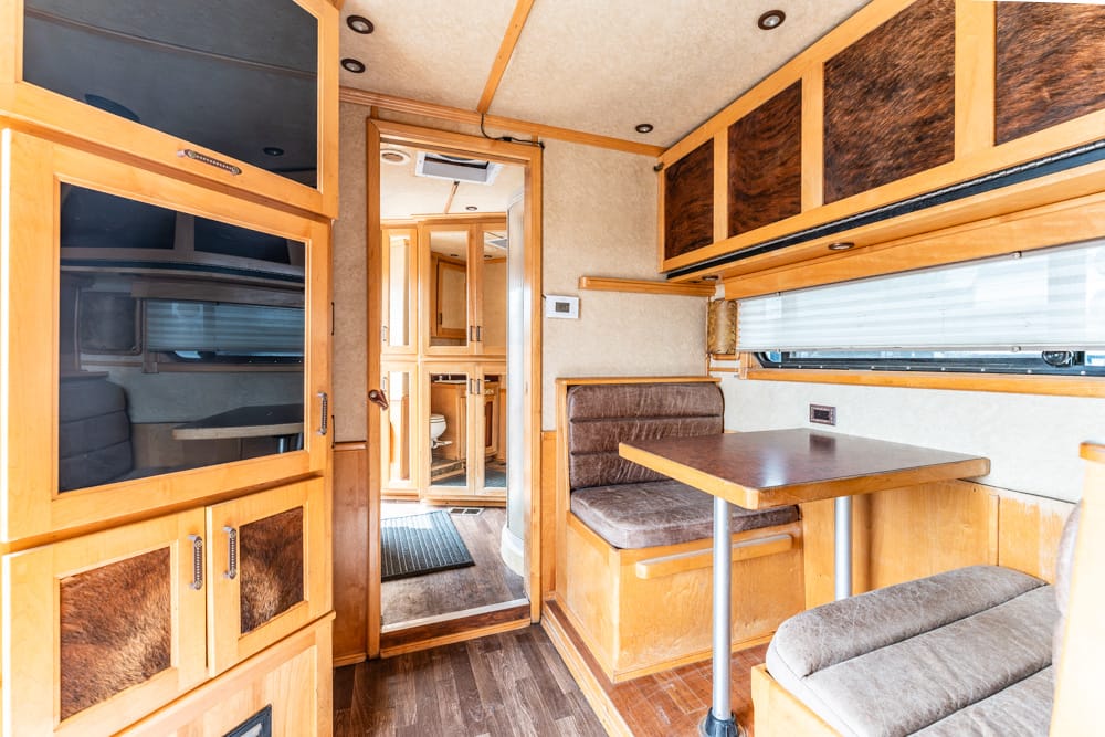 Compact RV interior with warm wood cabinetry, a small dinette with brown cushions, and overhead storage, seen toward a doorway to the back area.
