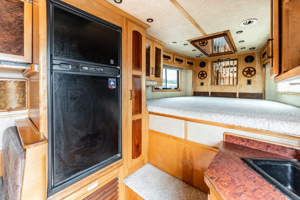 Wood-paneled RV interior with a tall black fridge beside cabinetry and a raised bed area.