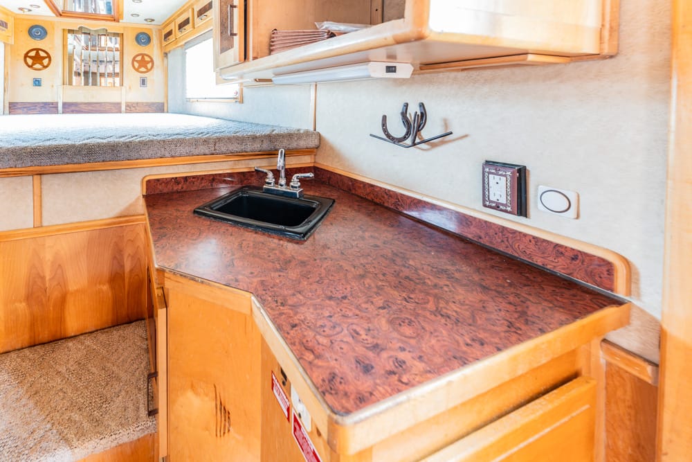 Compact boat galley with marbled brown countertop, black sink, and wooden cabinets; bunk area visible in background.