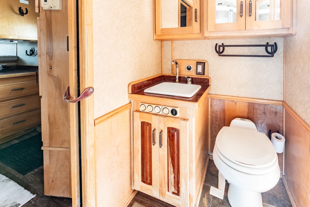 RV bathroom with wooden cabinet vanity, sink, and mirrored medicine cabinet; toilet to the right with a spare toilet roll nearby.