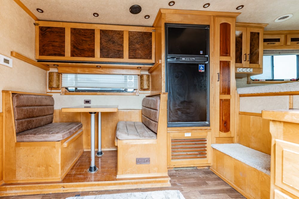 RV dining area with two brown cushioned benches around a small table, wood cabinetry overhead, and a tall black refrigerator in the background surrounding a window lighted kitchen space.