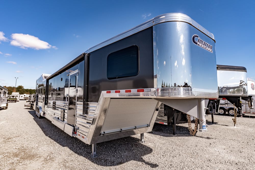 Large silver and black fifth-wheel RV trailer parked on a gravel lot under a clear blue sky, reflection visible on its shiny sides.