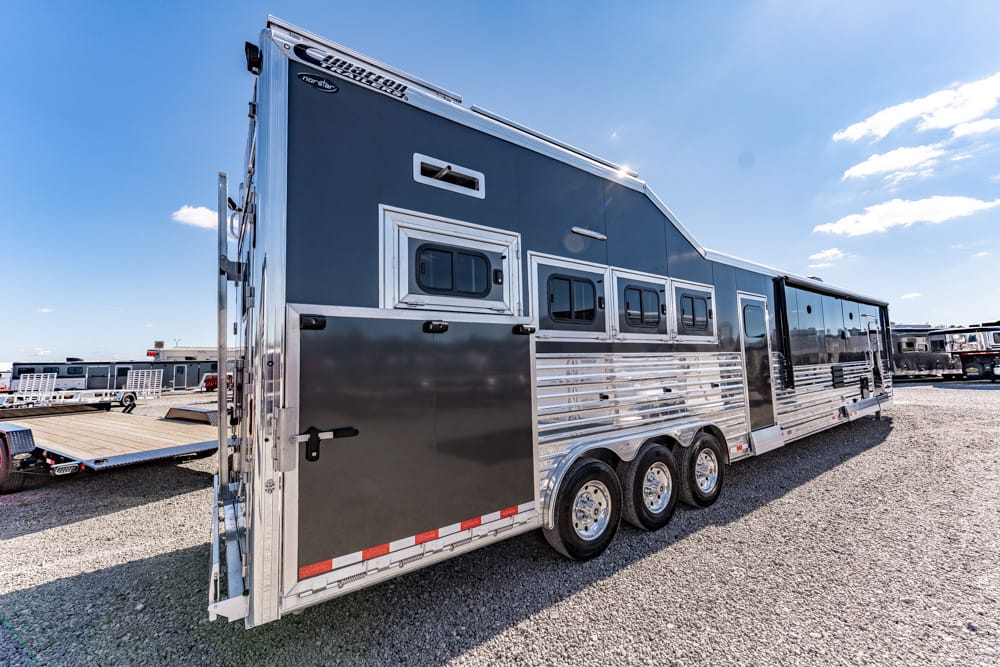 Long aluminum horse trailer with a dark upper half and multiple windows parked on a gravel lot under a blue sky.