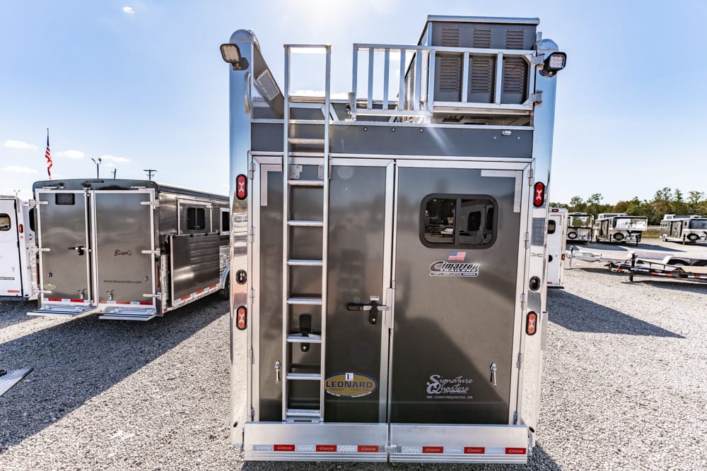 Rear view of a silver utility trailer with a side ladder, storage doors, and branding stickers, parked on gravel with other trailers nearby.