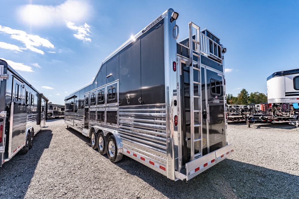 Shiny aluminum horse trailer with three axles parked in a gravel lot, with other trailers in the background on a sunny day.