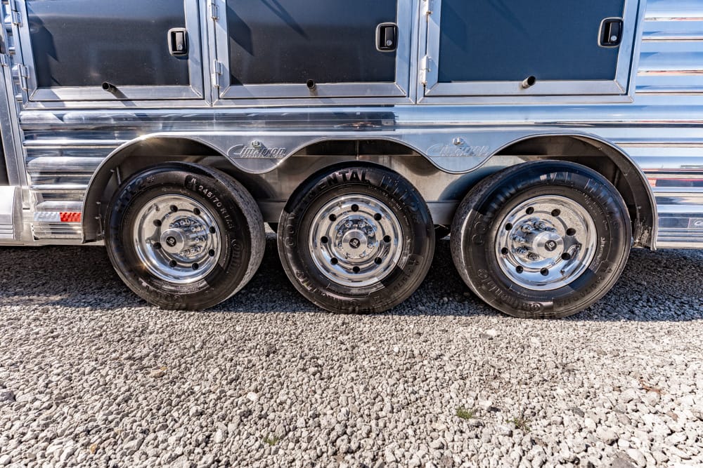 Close-up of a three-wheel axle on a shiny silver trailer, showing chrome rims and black tires on a gravel surface