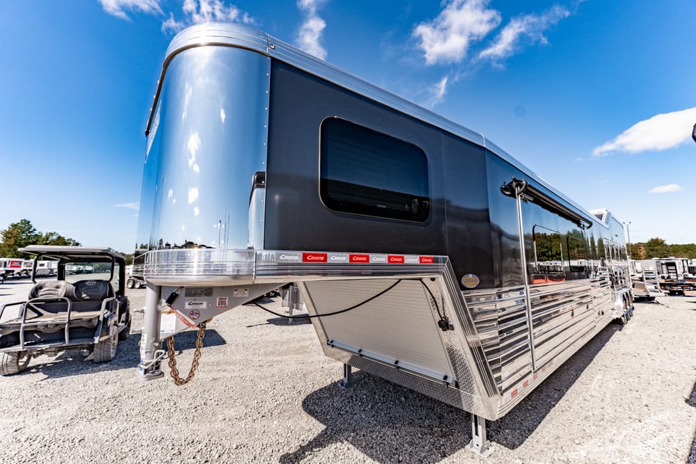Large chrome travel trailer with blue siding parked on a gravel lot; hitch and storage compartments visible under a blue sky.