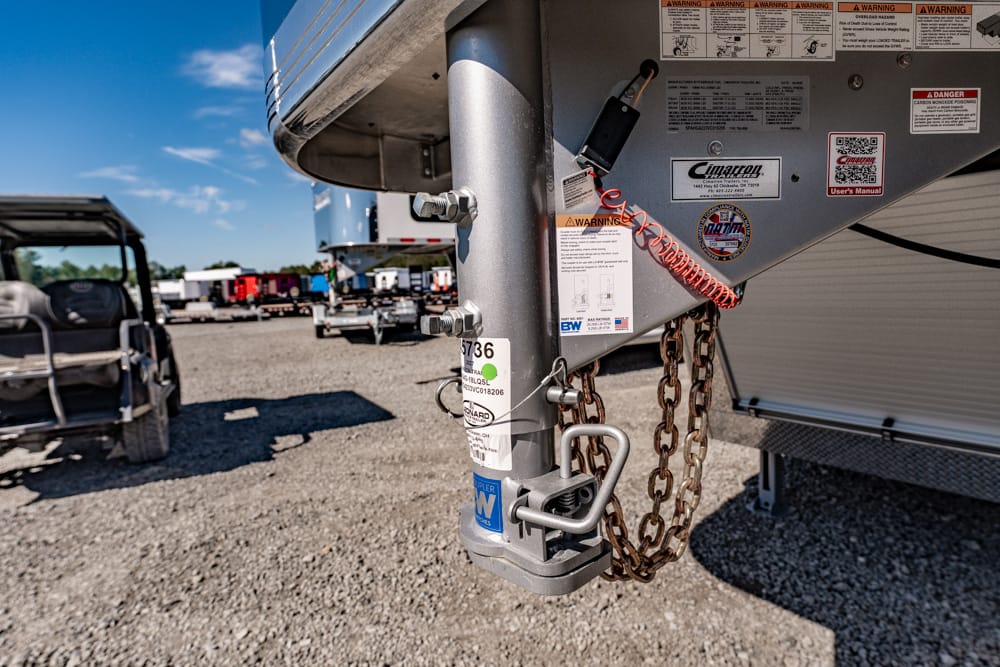 Close-up of a trailer hitch with safety chains and various warning stickers, on a gravel lot with other trailers in the background under a blue sky.