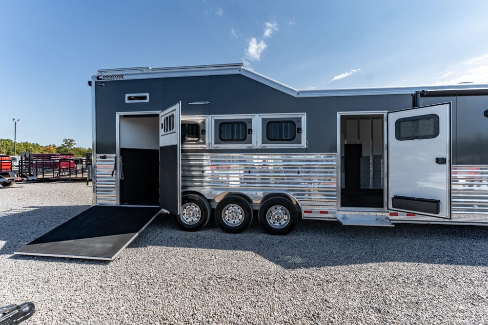 Long gray and chrome horse trailer with three axles, windows along the side, and a lowered ramp for loading horses on a gravel lot.
