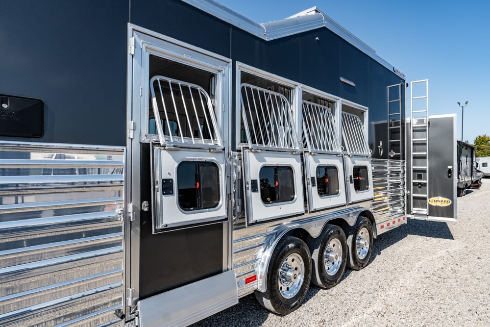 Side view of a dark horse trailer with three open stalls, barred windows, and shiny chrome trim on a gravel lot.
