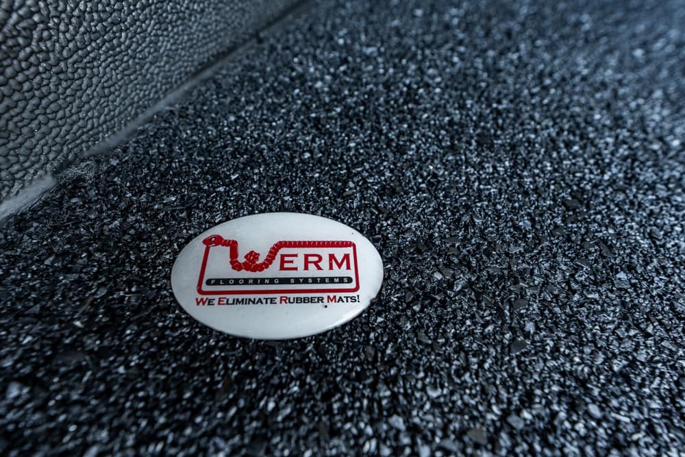 Close-up of a white oval badge on dark textured rubber flooring, reading 'We Eliminate Rubber Mats' with a red logo.